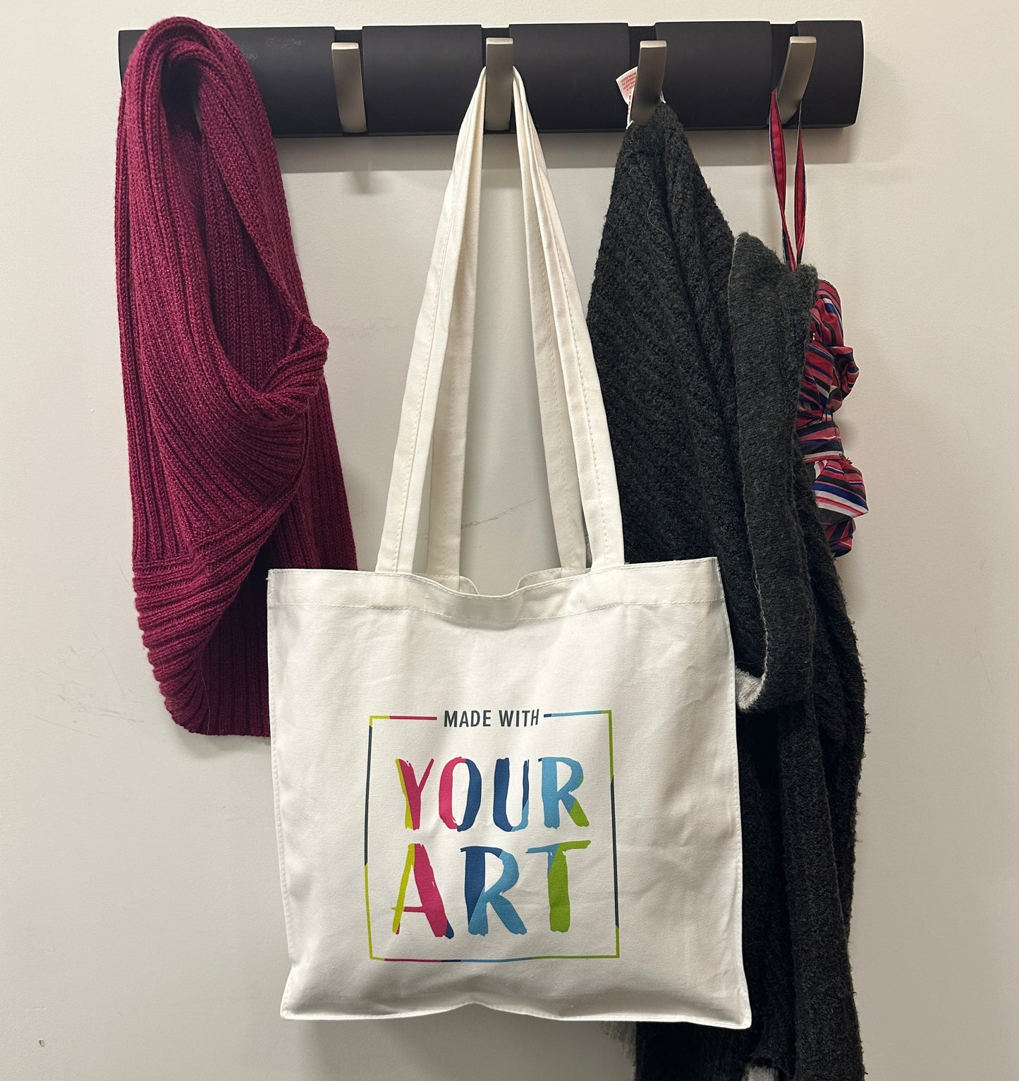Tote bag hanging on coat rack with logo Made With Your Art on it